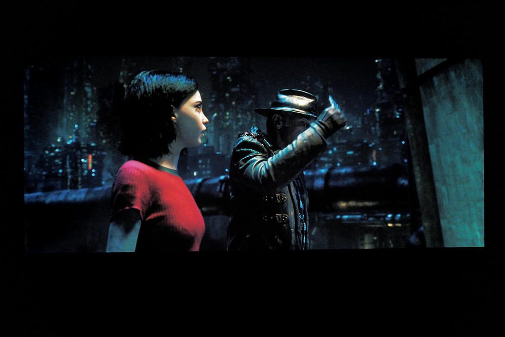 kobieta w czerwonej suknik, obok niej mężczyzna w ciemnym ubiorze - scena rozgrywa się na ciemnym tle