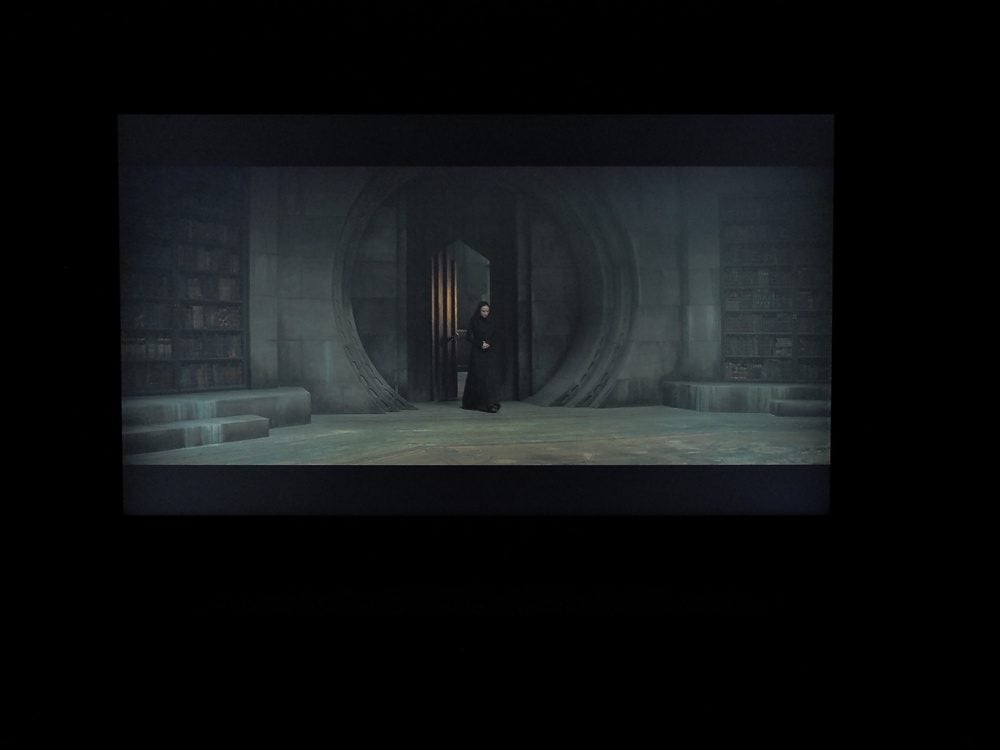 w centralnej części ekranu ogromne, okrągłe wrota, przechodzi przez nie kobieta ubrana w czarną suknię